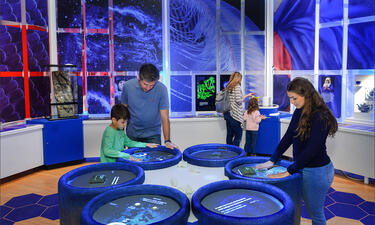 In the center of a blue room a family surrounds several circular digital stations that demonstrate the process of dyeing indigo fabric