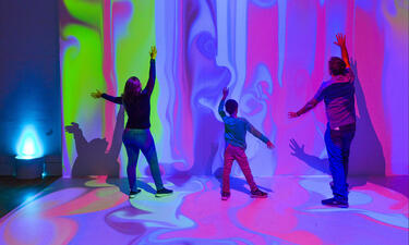 A woman, child and man with arms outstretched are seen from behind facing a wall displaying a projection of colorful paint dripping from ceiling
