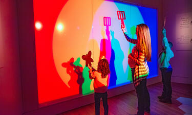 A woman and two children stand in front of a white wall, their shadows are shown in several different colors