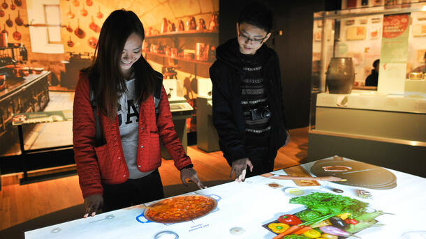 Two teenagers stand over an interactive table showing a projection of meals being cooked