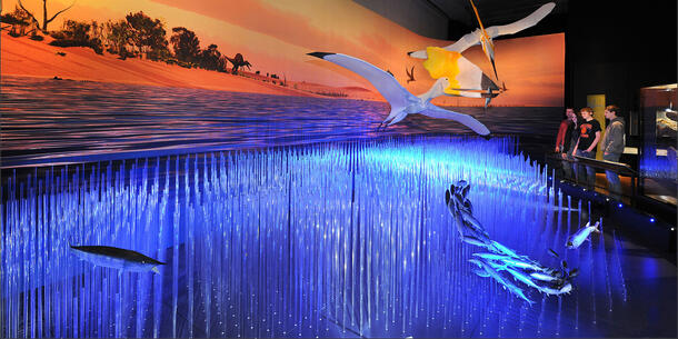 An exhibition hall with models of pterosaurs flying over a model of seashore water with fish. In the background is a sandy beach where walks a large dinosaur.