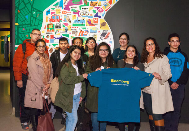 Group of teens and adults stands in front of the logo for the Museum's !Cuba! exhibition, two teens in front hold a t-shirt that says "Bloomberg".