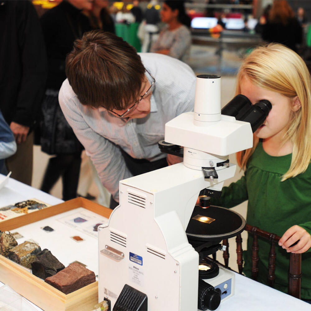 A child looks at a slide through a microscope while an adult watches over, with rock specimens on the table beside them.
