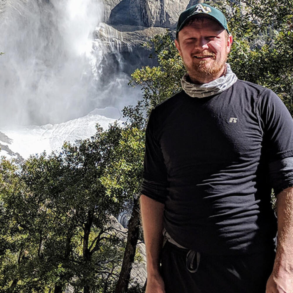 Erik Wright wears a baseball cap and poses for a smiling portrait outdoors, with a waterfall in view in the background.