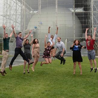 Nine students in the MAT Program jump in the air on the lawn in front of the Rose Center for Earth and Space.