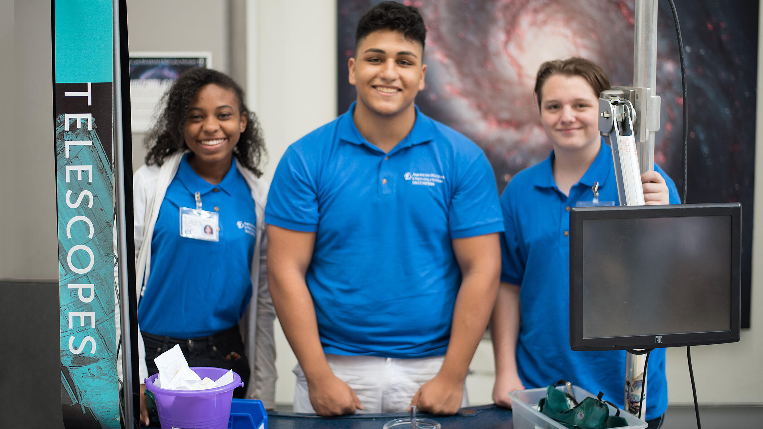 Three teenaged students stand behind a table with a computer screen. A vertical banner with text that says "Telescopes" is next to them on the left. 