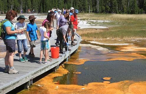 adults and children stand on a short pier and watch a park ranger collect a water sample from a wetland