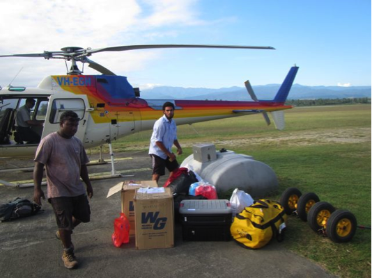 Two men on a grassy landing field with supplies and equipment in the foreground a helicopter in the background.