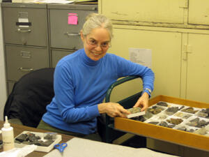 A woman smiling at the camera and seated next to a specimen tray containing multiple artifacts.