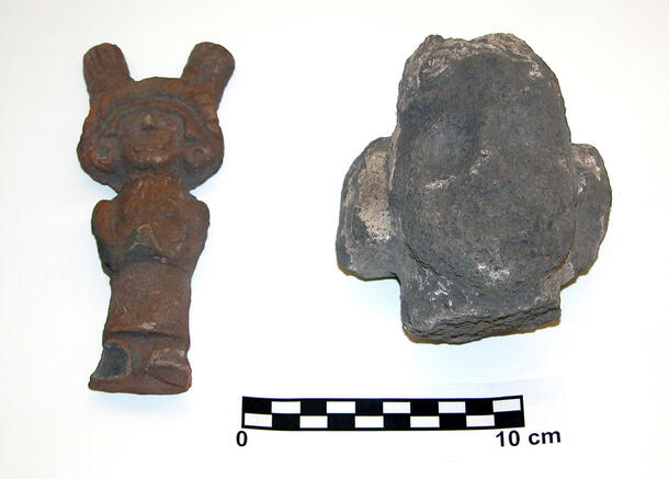 Two small idols, one in the shape of a human head broken off from a larger object, the other a human figure wearing a large headdress.