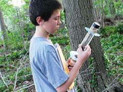 A boy standing in a forest beside a tree trunk holding a cylindrical piece of equipment.