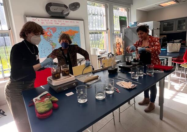 Two participants stand by the workshop leader at a lab bench wearing blue latex gloves, tending to beakers full of dye, stirring a brown liquid. 