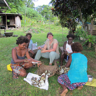 Five people sit on grass around baskets full of shells as they shuck and prepare the shells and invertebrates for cooking. 