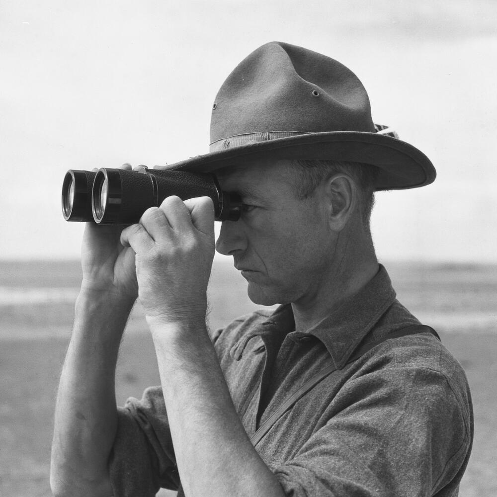 Archival photograph of a person in a wide brim hat looking through a pair of binoculars.