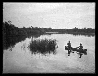 Wide shot of two people canoeing on flat still water. A tuft of marsh grass growing out of the water near canoe. Low scrub surrounds water.