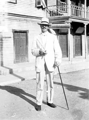 A man, Marshal H. Saville, in a suit, hat, and walking stick, standing in front of a building.