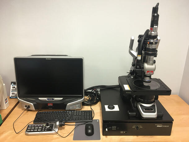 A computer (with keyboard, mouse, and joystick) on a wooden desk, to the left of a digital microscope.