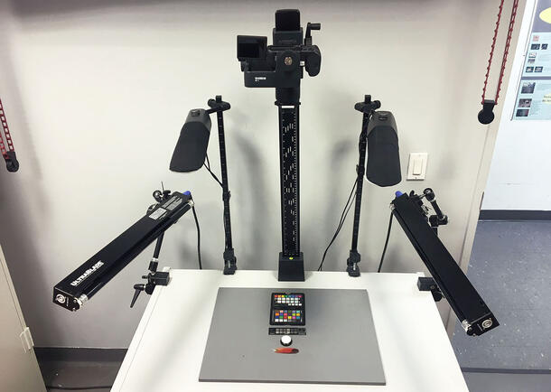 A copy-stand with four lamps on articulating supports and a central vertical column camera mount. One color target, one UV-imaging target, one white circular reflectance standard, and one feather are on the copy stand.