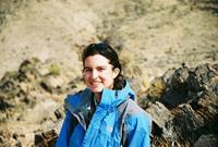 Outdoor photo of a woman, Melanie Hopkins, smiling at the camera, wearing a blue windbreaker, with a dry rocky surface in the background.