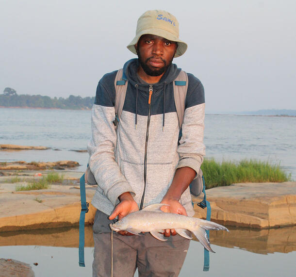 Tobit Liyandja stands near a body of water holding a fish specimen in both hands.