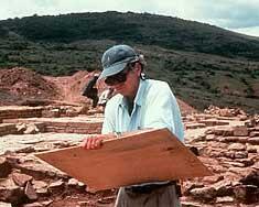 A man at an excavation site wearing a baseball cap and sunglasses holding a wood board.