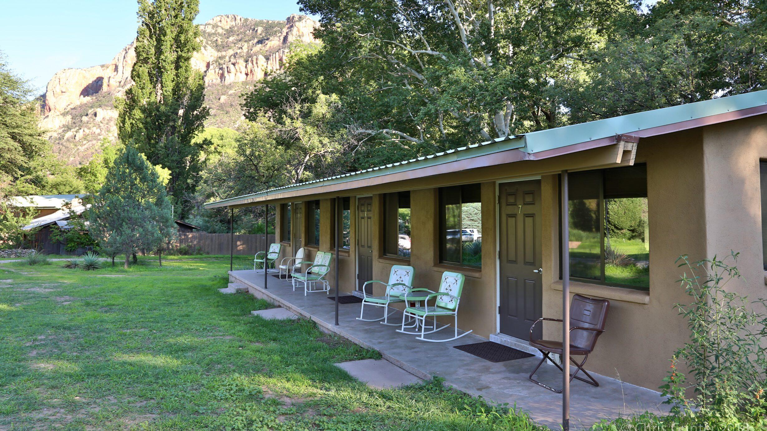 Porch with metal chairs in front of lodging rooms with trees and mountains in the background