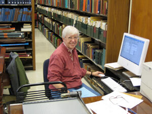 A woman smiling at the camera and seated at a desk with a computer, with library shelves full of books in the background.