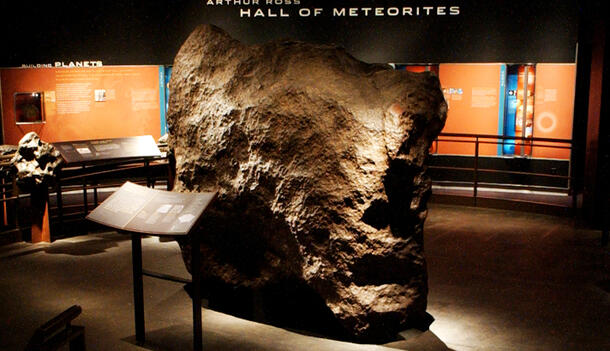 A large, reddish-black iron meteorite situated next to a label deck in the Hall of Meteorites.