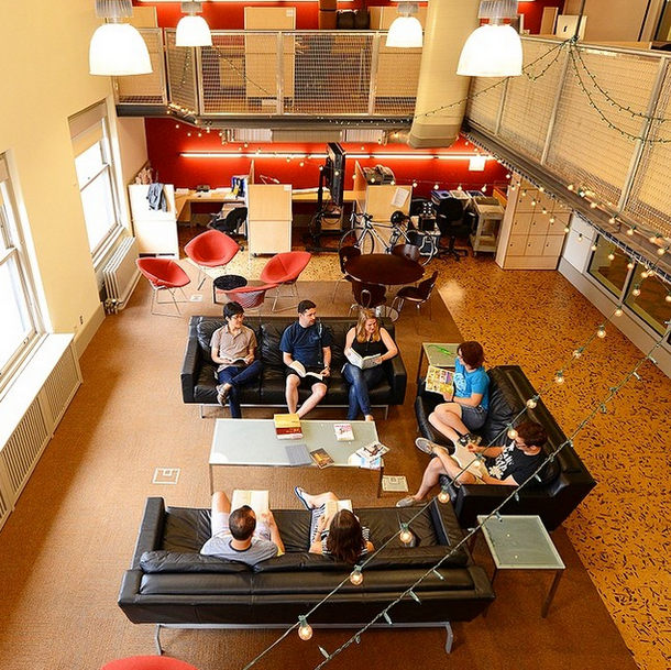 Seven people, students at the Museum’s Richard Gilder Graduate School, sitting on three sofas, with books on their laps, in a large room with windows, desks, tables and chairs.