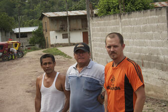Three men outdoors dressed in T-shirts posing for the camera with a cinderblock wall and two low buildings in the background.