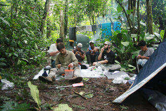 Six people sitting on the damp brown soil of a campsite in a lush green forest.