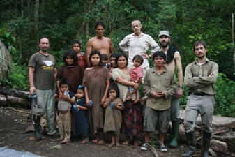 Sixteen people, men, women, and children, standing posing for the camera in a lush green forest clearing in front of a stone wall