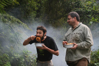 Two men in a lush green forest clearing posing for the camera while eating food from their two bowls.