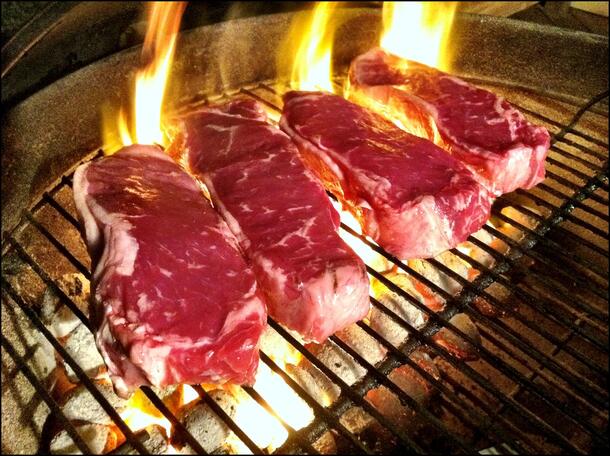 Four raw red meat steaks cooking on a grill with flames coming up beneath them.