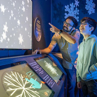 Two Museum visitors, an adult and teenager, watch a digital display of snowflakes in front of an interactive panel about snowflakes.