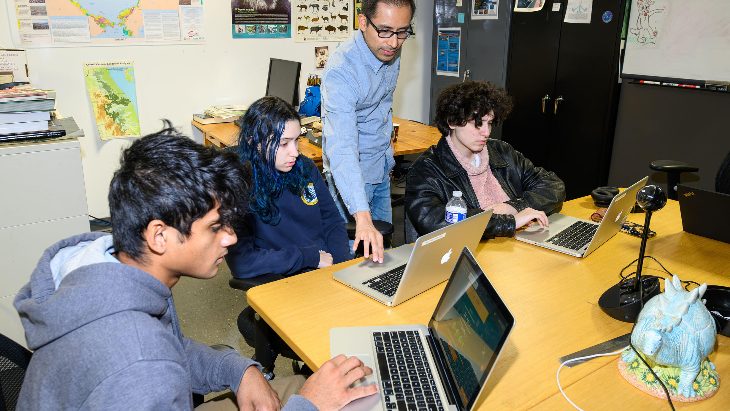 Three students from the Science Research Mentoring Program sit at a large desk on their laptops as an instructor helps one of them.
