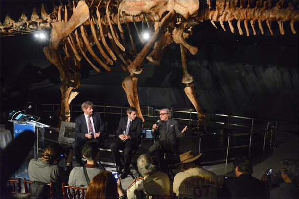 Paleontologists Mike Novacek, Diego Pol, and Mark Norell seated beneath the Titanosaur fossil, holding microphones in front of a seated audience.