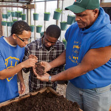 A tall Growing Power member put out their hands which are holding dirt and worms, while two other people look at the worms.