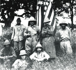 Theodore Roosevelt (standing, second from left) and six other people sit and stand in front of American flag and trees.