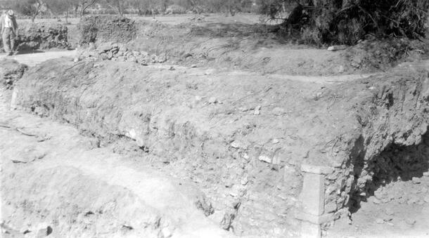 An excavation area with dry soil showing a partially exposed wall, and a person standing in the background. The photo caption states: "West platform looking northeast."