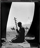 In silhouette, a person kneeling in a tent while holding a long object. In the background are two people astride camels, with hills in the distance.