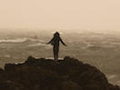 Person standing on a cliff with arms down and outstretched, silhouetted against sky.