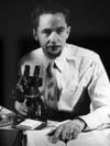 Man wearing shirt and tie sitting at table with microscope, looking at camera: Mont A. Cazier, former curator at the American Museum of Natural History, as a young man.