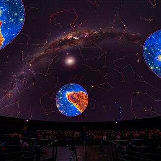 An audience looks up at three bright, splotchy depiction of the Milky Way inside the Hayden Planetarium.