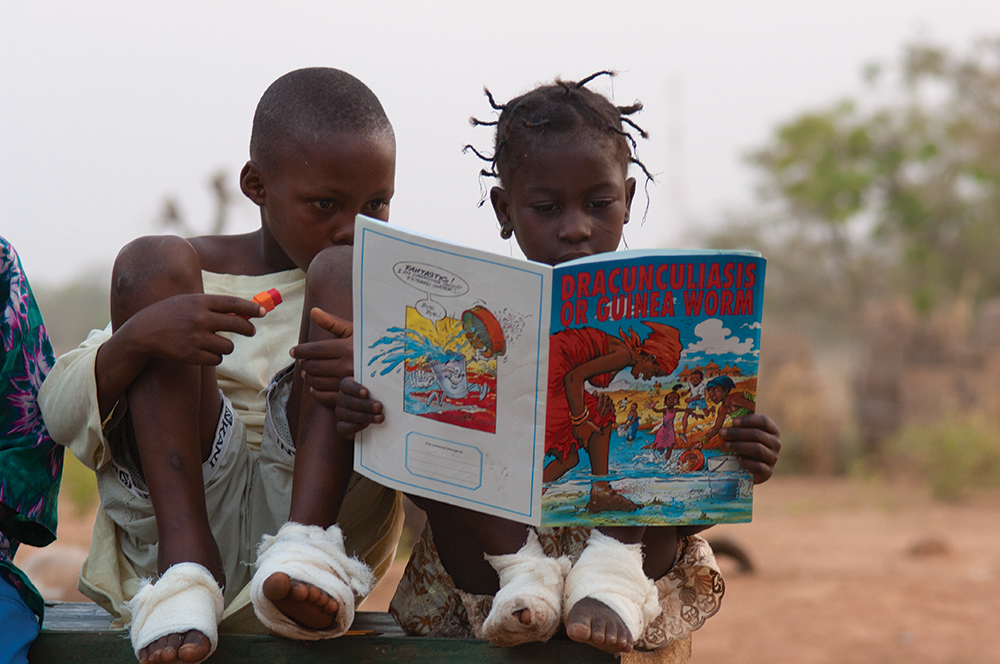 Two children, each with both feet wrapped in bandages, reading a book titled, "Dracunculiasis or Guinea Worm."