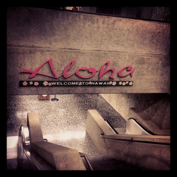 A photo from the top of an escalator shows two escalators and a staircase leading to a black marble spotted floor. The wall above has the words “Aloha” and “Welcome to Hawaii.