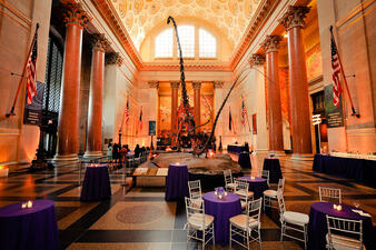 Small round tables with four chairs each surround a centrally-displayed tall fossil dinosaur mount in the Museum's rotunda.
