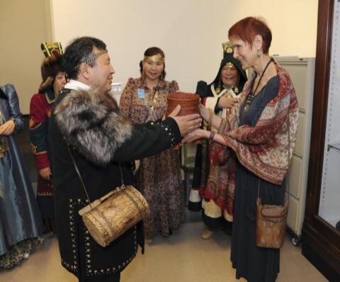 Laurel Kendall, the Museum’s Curator of Asian Ethnology accepting a large handmade wood goblet from Sakha delegation member Fedor Chairin as other members, all dressed in cultural attire, watch.