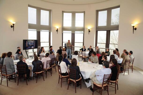 People seated at five luncheon tables set up in the Museum’s Astor Turret, listening to a man standing and giving a presentation.