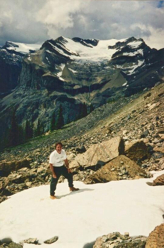 Martin Shugar stands on a snowy patch on a rocky mountainside, with the peaks of another lightly snow topped mountain in the background.